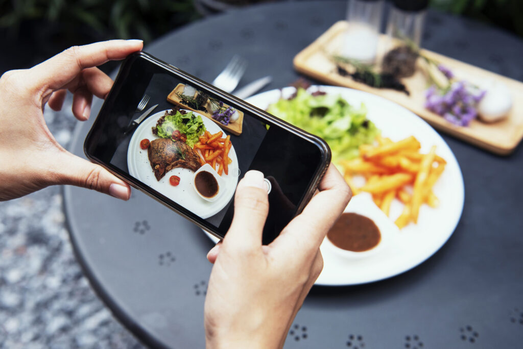 blogger hands with smartphone photographing pork chop steak with vegetable salad and potato chips on white plate in the garden. top view.